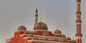 A striking red-brick mosque in Al Barsha, Dubai, with intricate Islamic calligraphy, multiple domes with golden details, and tall, ornate minarets, standing against a hazy sky