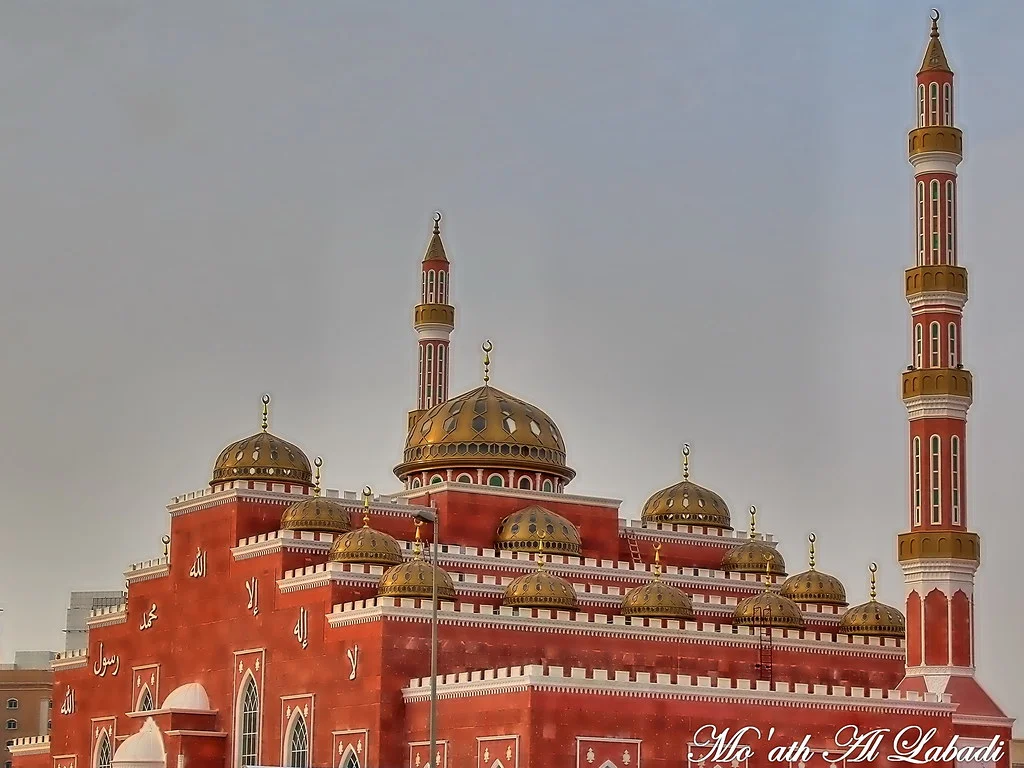 A striking red-brick mosque in Al Barsha, Dubai, with intricate Islamic calligraphy, multiple domes with golden details, and tall, ornate minarets, standing against a hazy sky